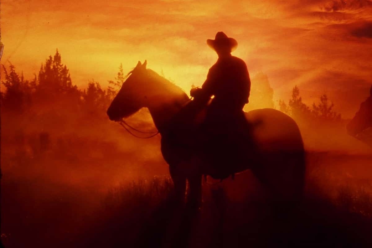 Man on horseback wearing cowboy hat with red sunset behind him.