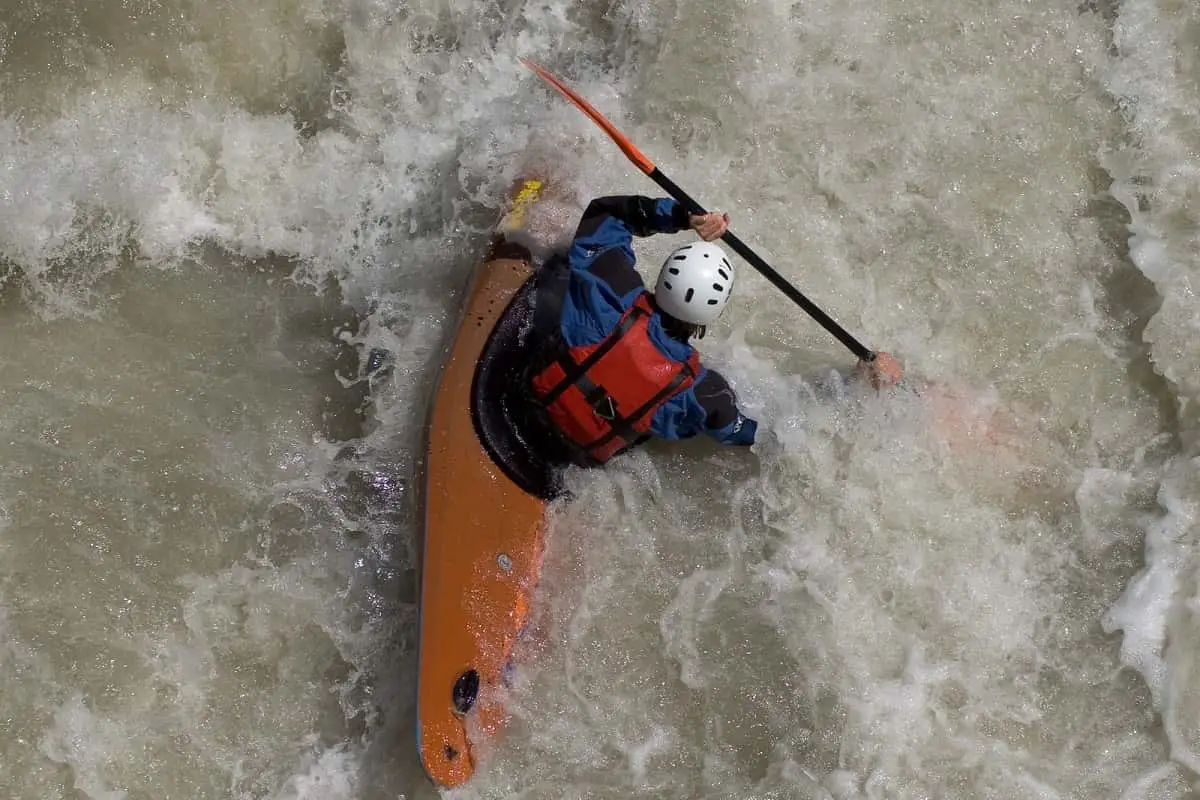 Person wearing white helmet and red safety vest kayaking in white water rapids