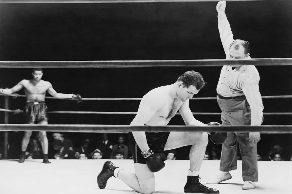 black and white image of boxing match with referee giving a standing count while the other boxer waits in his corner
