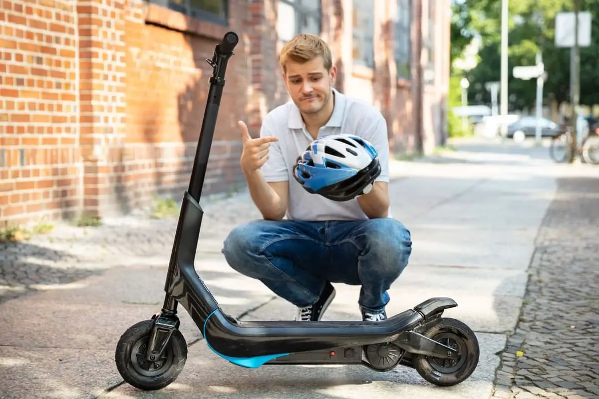 man crouching beside electric scooter pointing at his bicycle helmet