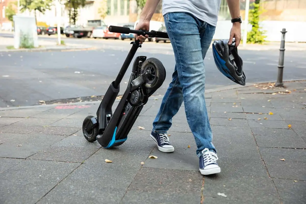 man pulling folded electric scooter while holding his bicycle helmet