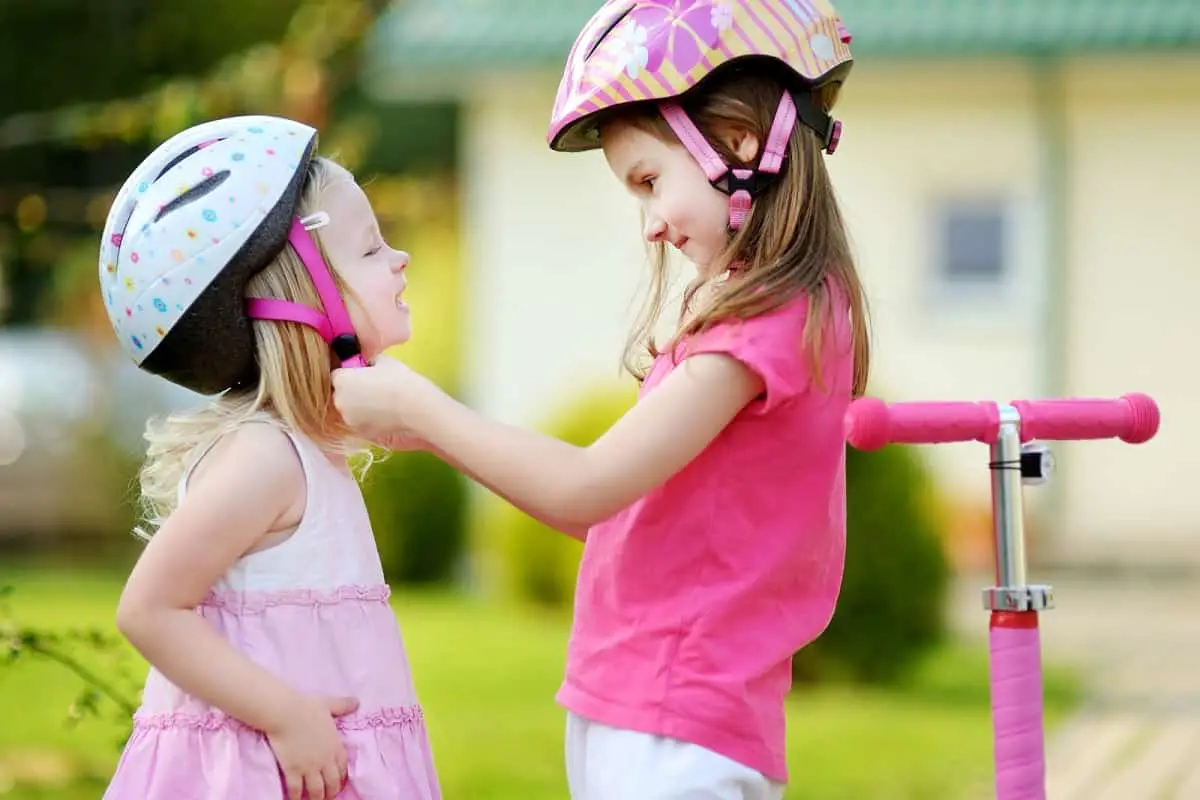 older girl wearing bike helmet helps younger child to fasten helmet chinstrap