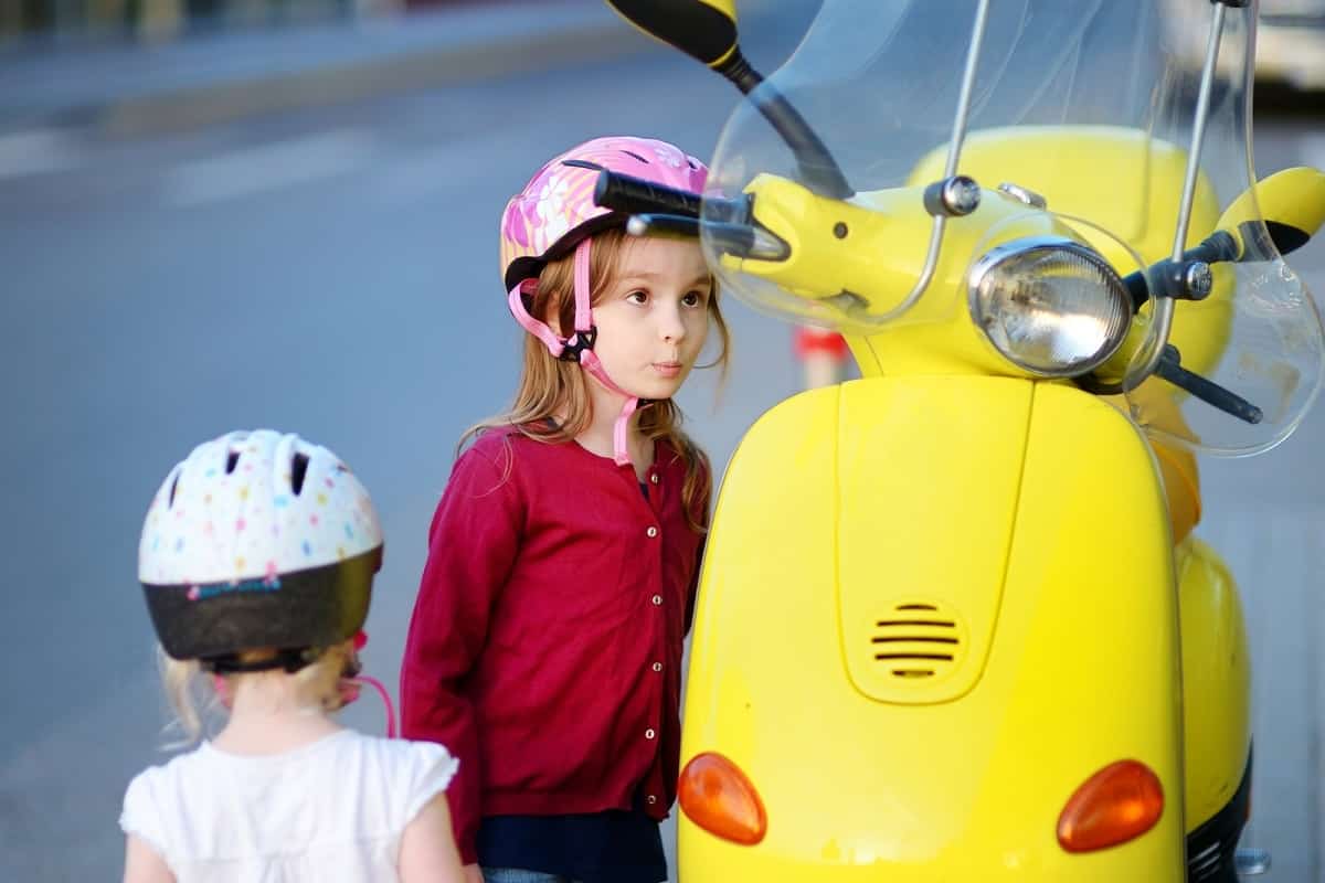 2 kids wearing bike helmets looking at a moped