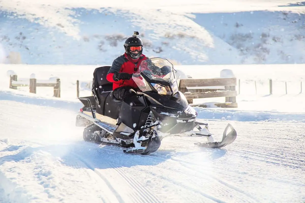 man wearing black helmet with gopro attached riding snowmobile along snowy road with wooden fence alongside