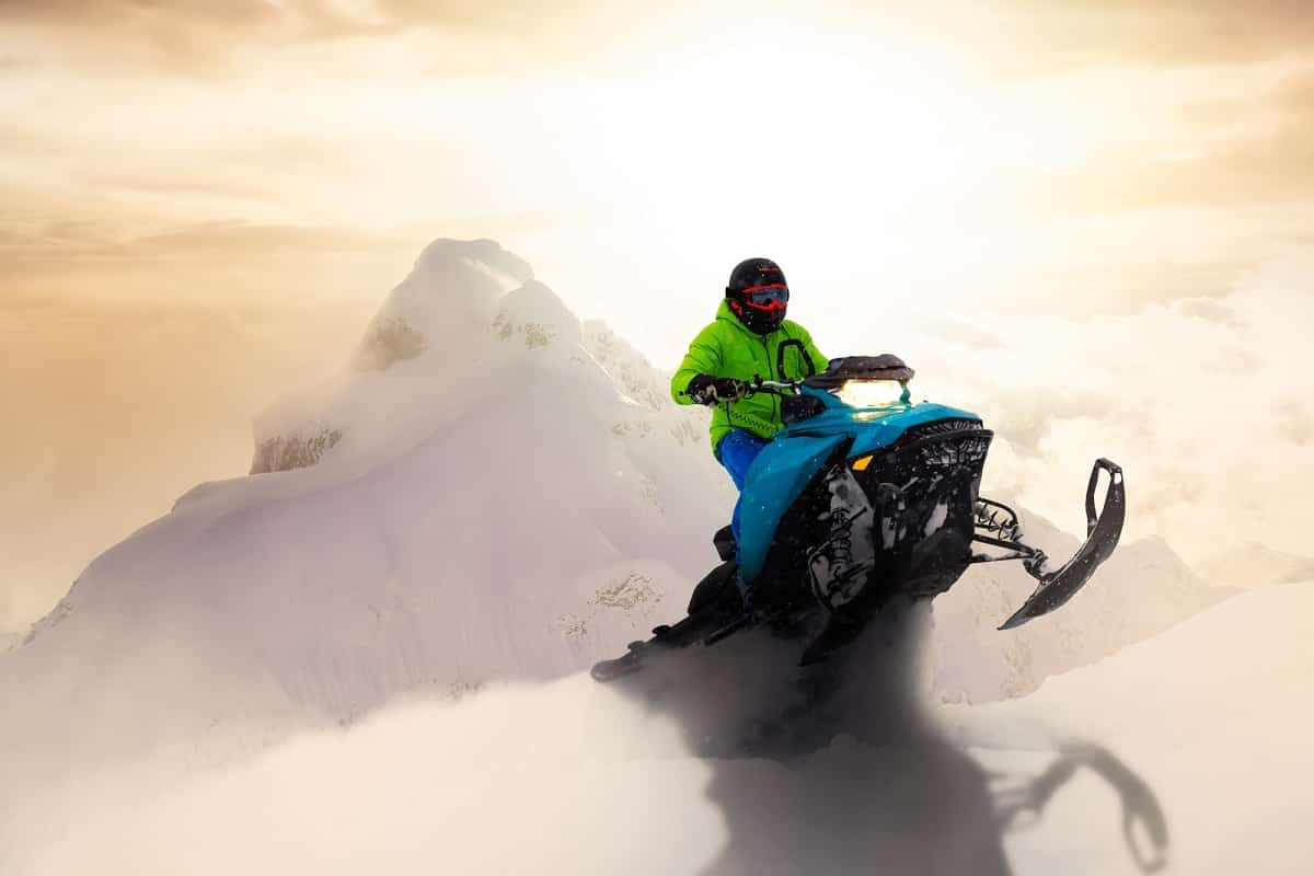 man wearing green jacket and black helmet rides snowmobile over jump in powdery snow