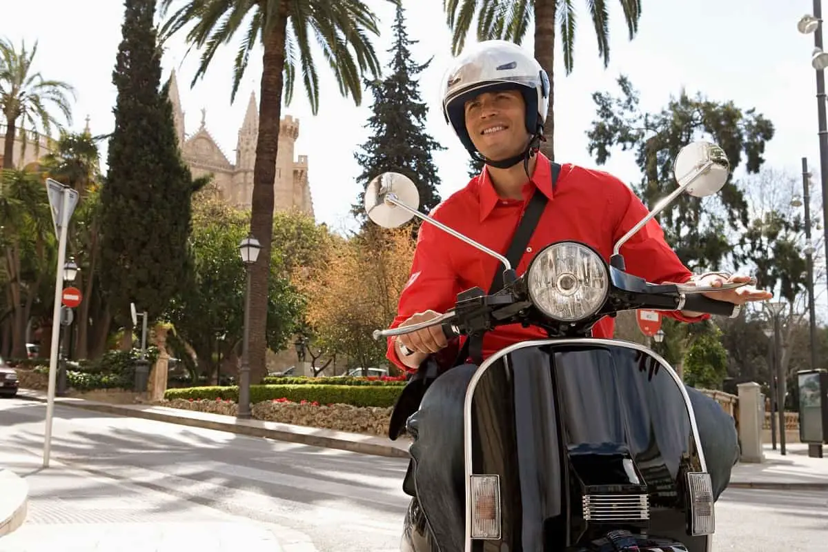 man in red shirt wearing a white helmet riding a moped in the city near a garden