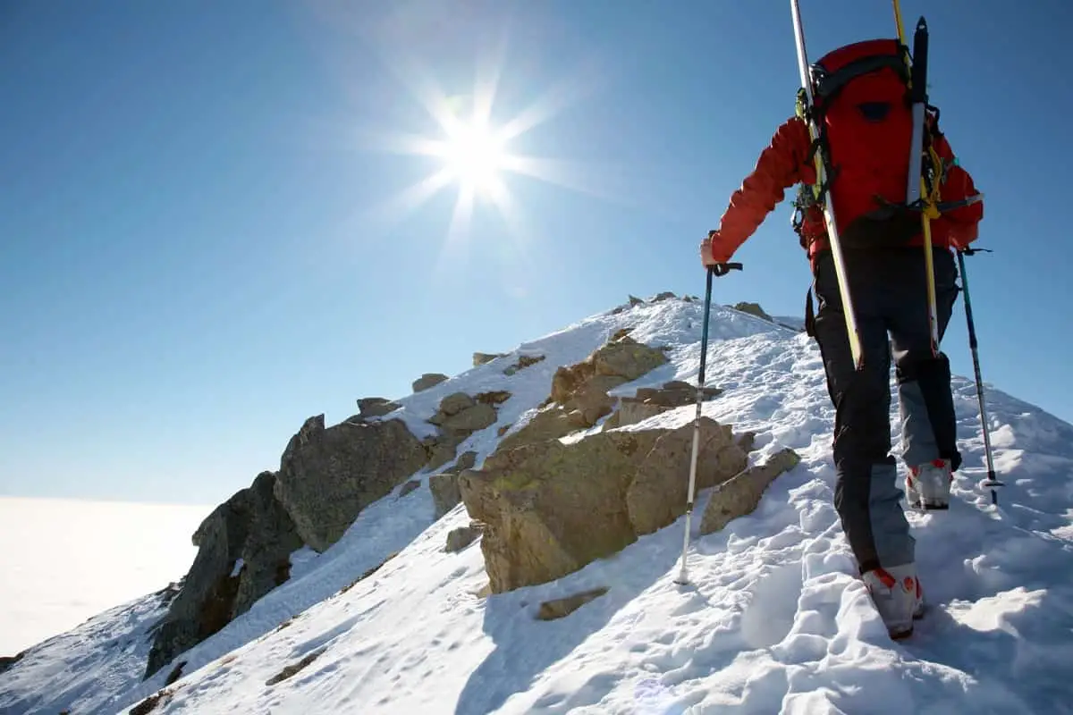 man in red jacket walking up snow covered mountain with snow skis strapped to his back