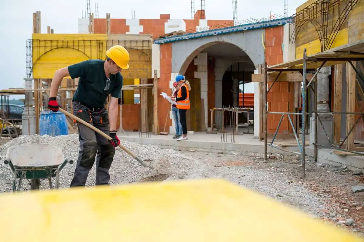 construction site with one worker shoveling gravel into a wheelbarrow while two engineers check the plan