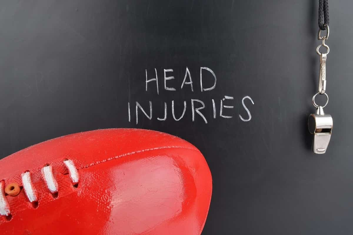 the words head injuries written on a chalk board in white chalk with a umpires whistle hanging on the right side and a red football on the left foreground