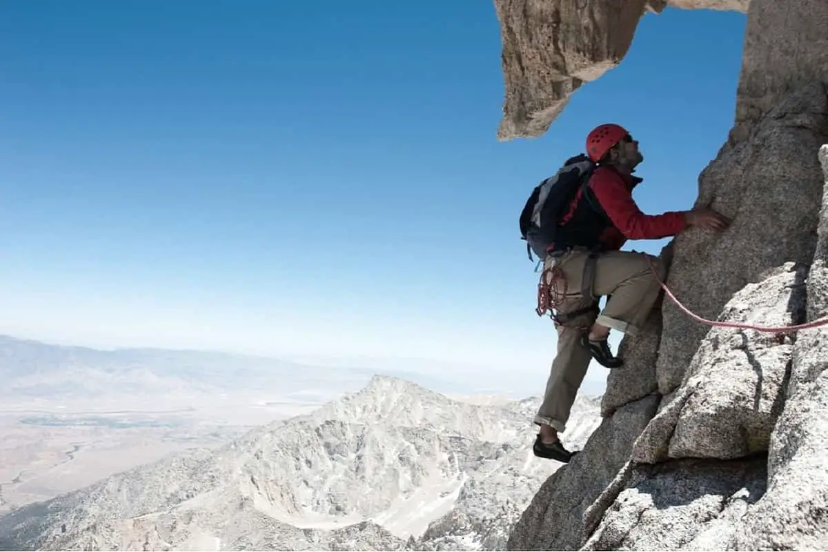 mountain climber wearing orange helmet and carrying a backpack on the side of a rocky mountain side