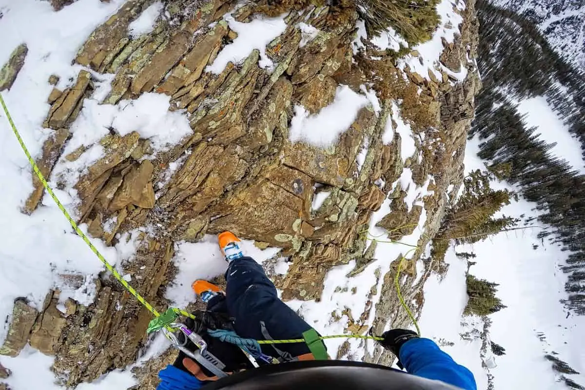 helmet-cam view of mountaineer looking down at his feet while standing on the side of a snowy mountain with snow and pine trees down below