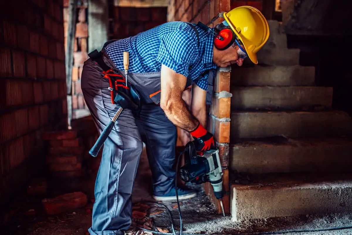 middle-aged house builder wearing yellow hard hat and ear protection drilling a hole in cement
