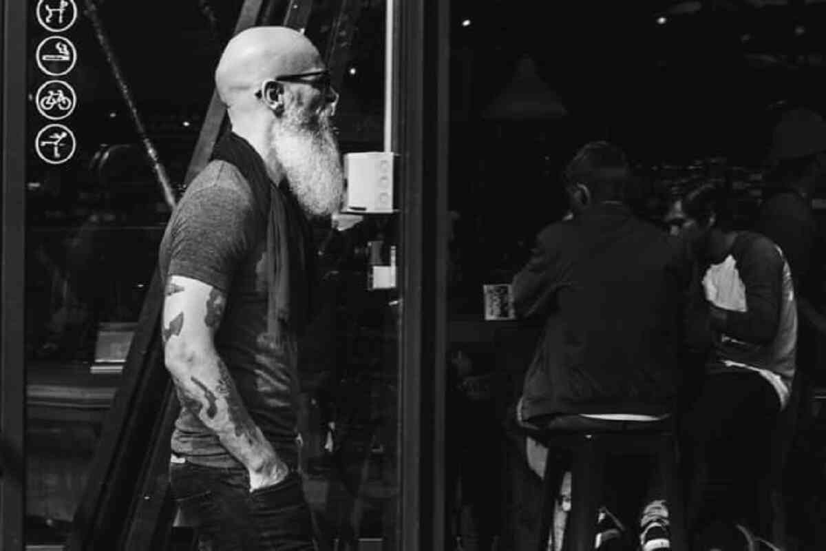 Do motorcycle helmets cause hair loss? Probably not.
Bald headed man with white beard and tattoos walking past a bar.