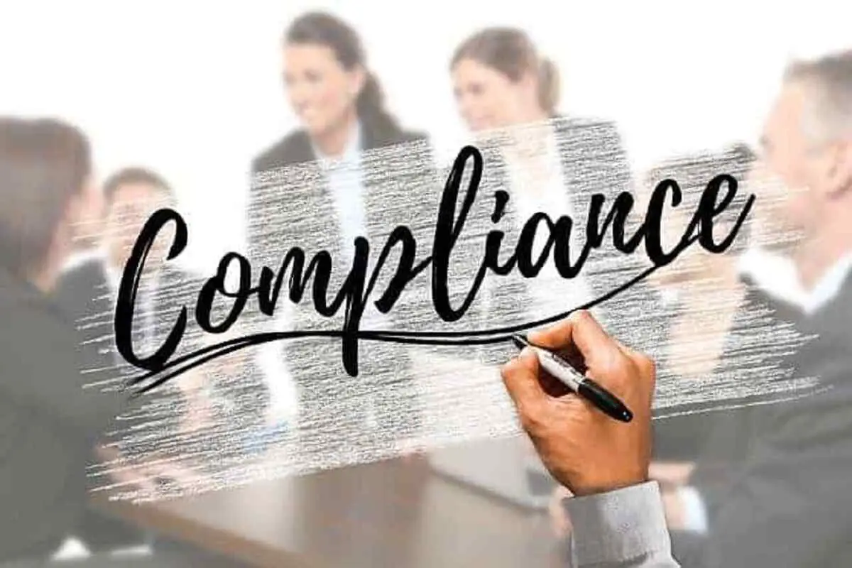 The word compliance written in black across a smoked glass window with a group of people on the other side of the glass