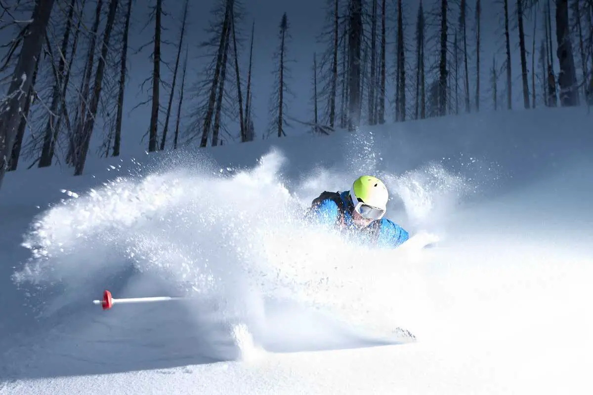 Skier wearing light blue and a yellow helmet skiing through fine powdery snow.