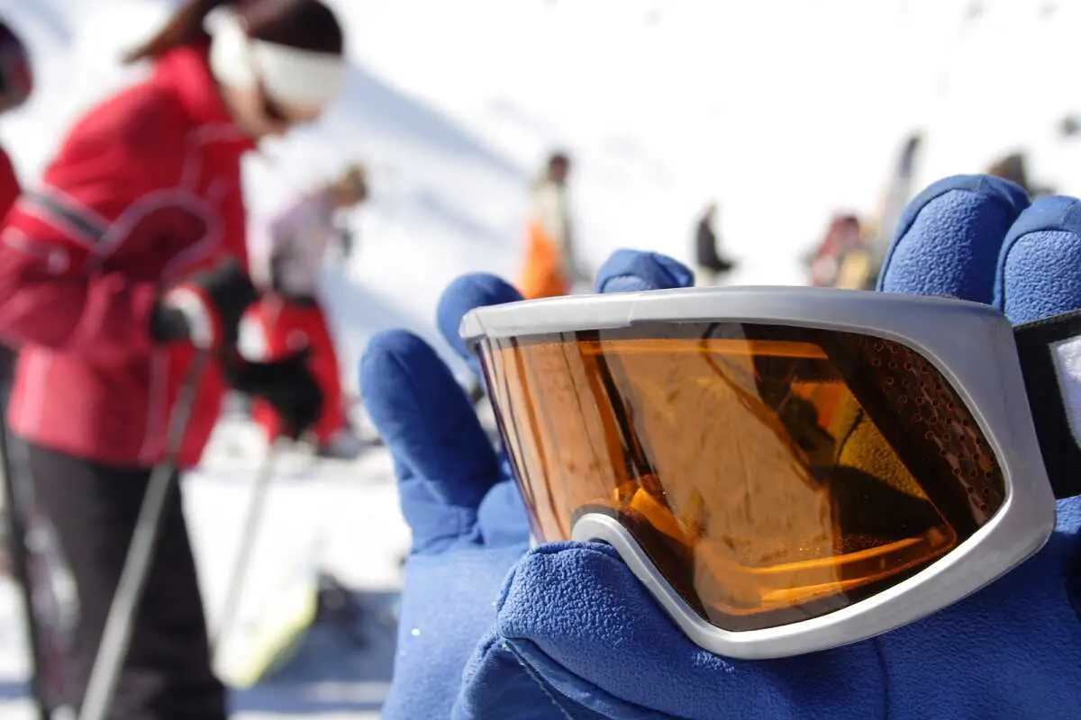 Blue gloved hand holding a grey framed pair of ski goggles with an orange lens.