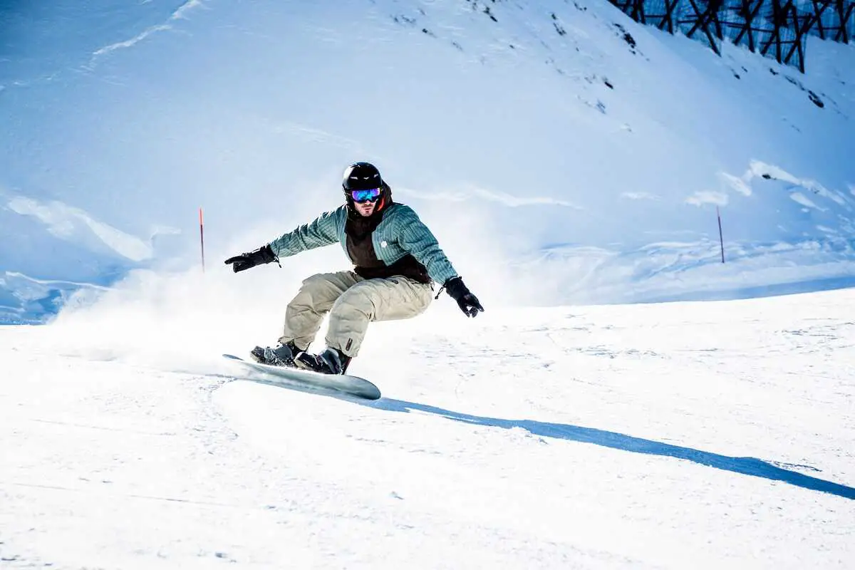 snowboarder wearing black ski helmet and light colored clothes with arms outstretched snowboarding along a slight ski slope.