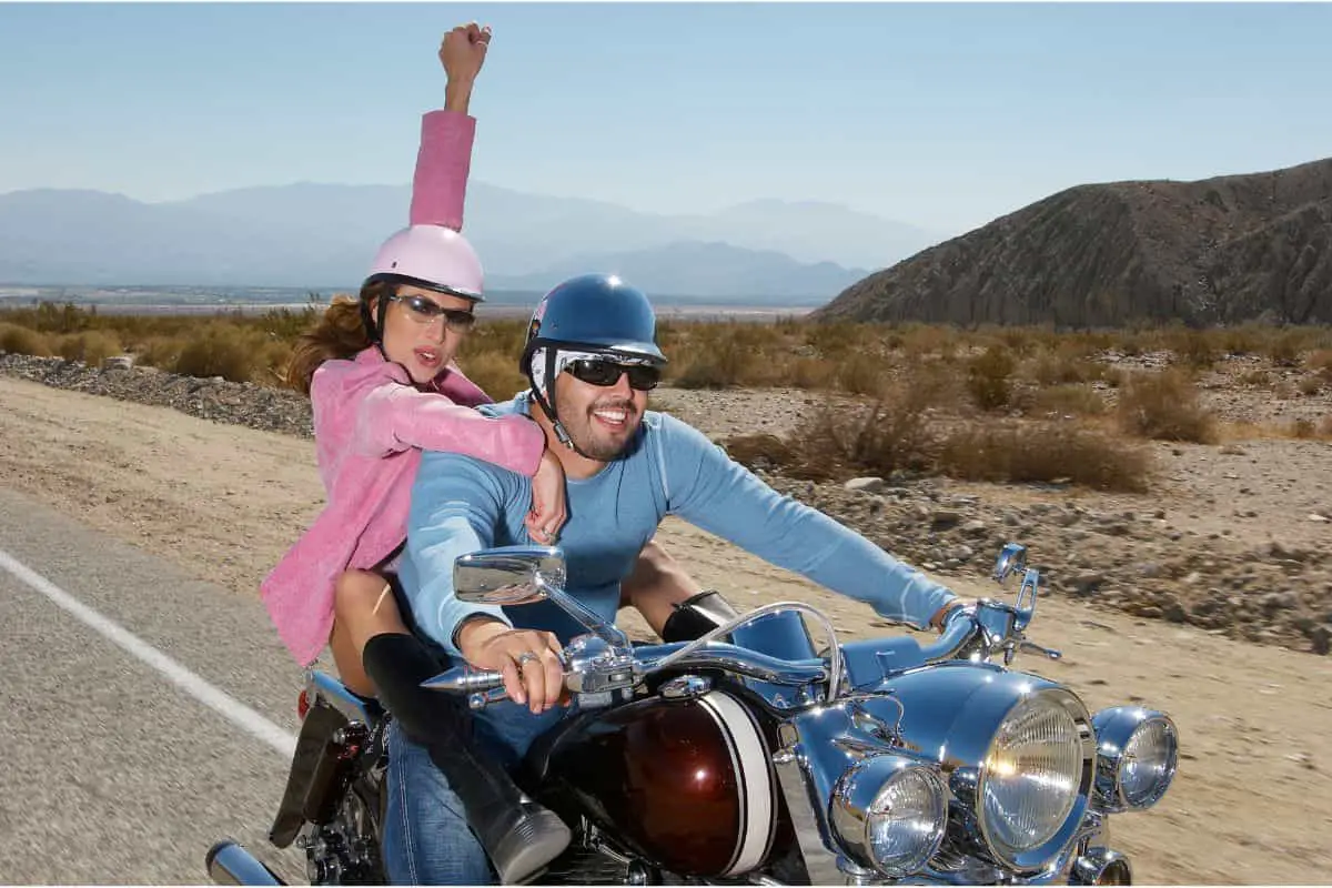 A nan and a woman wearing half helmets riding a motorcycle in the desert.