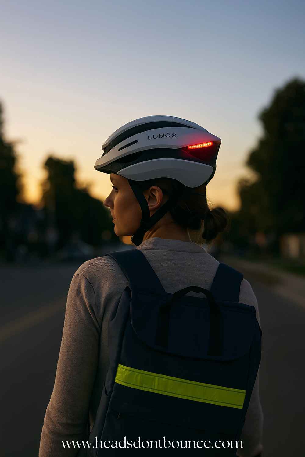 A young woman walking home at dusk wearing a white Lumos Ultra Smart bike helmet. The helmet's rear LED lights are illuminated.