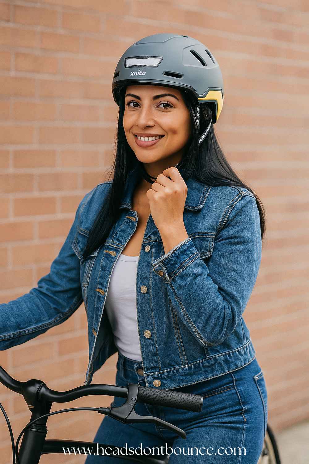 An attractive woman sitting on her bicycle wearing all denim and a XNITO urban bike helmet. Behind her is a brick wall.