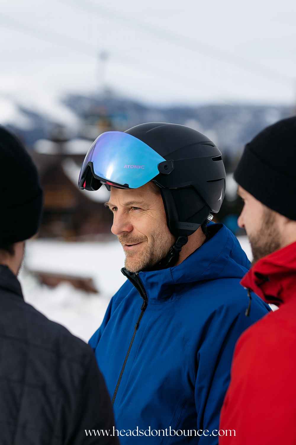 Forty-year-old male skier wearing a black visor helmet chatting with two friends at a snowy resort; chalet and lift structures visible in the background.