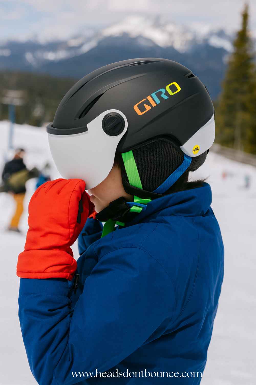 Photo of a young boy skier from the side adjusting his helmet visor while wearing a black Giro Buzz visor helmet with white lens and multicolor GIRO logo; skiers and mountains in the background.