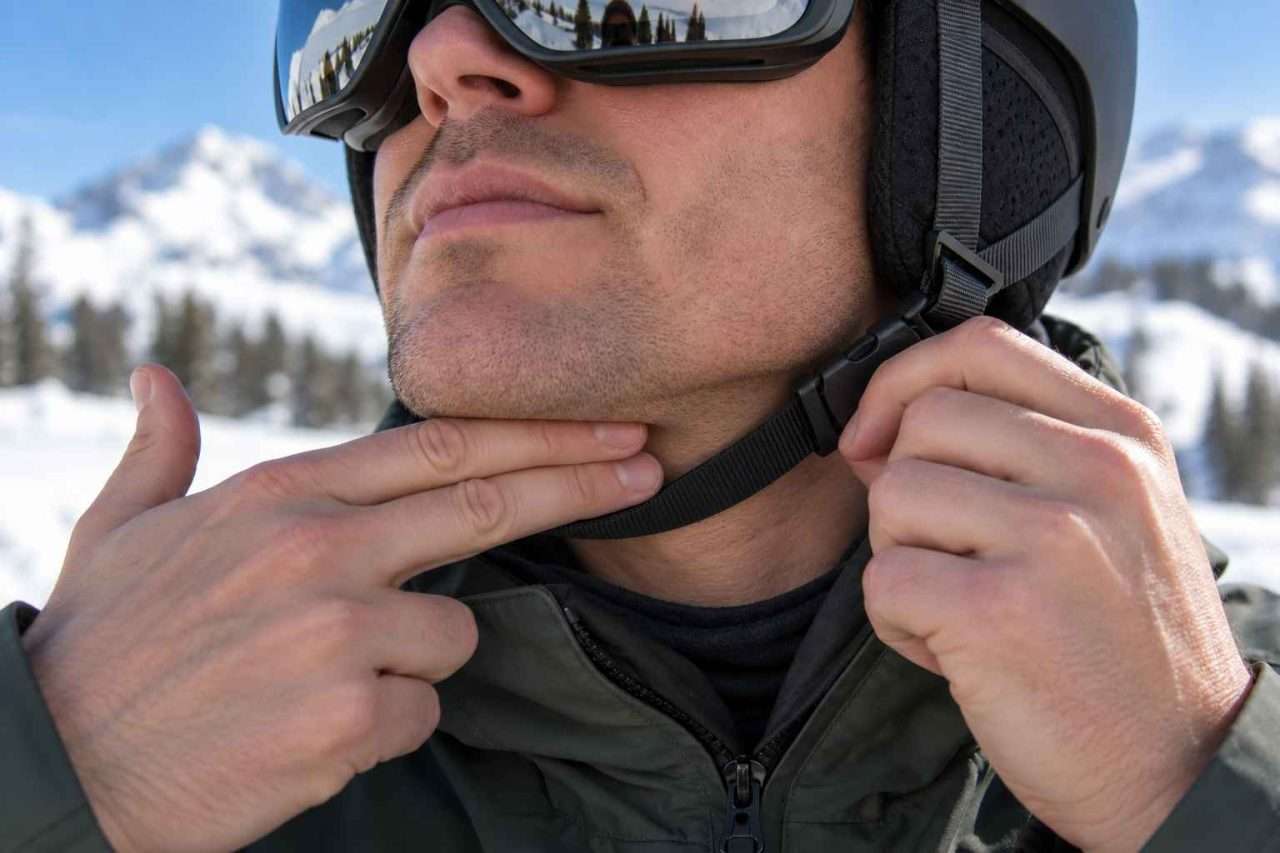 Skier checking ski helmet chin strap fit with two fingers between the black strap and the underside of his chin.