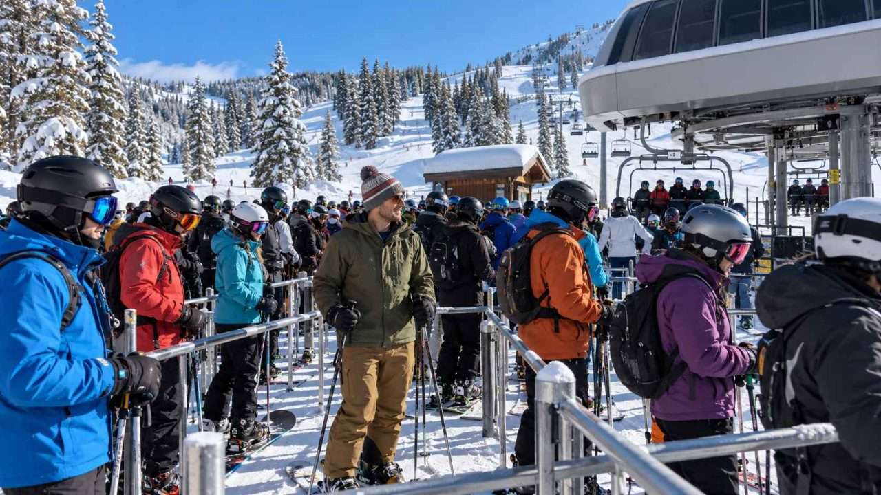 Skiers wearing ski helmets wait in a busy chairlift line, with one skier in a knit beanie among them.